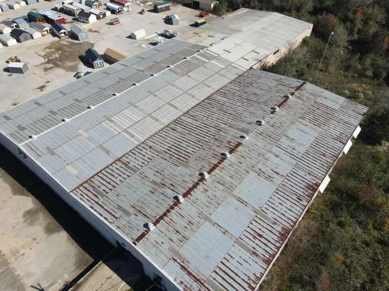 Aerial view of a large industrial building with a corrugated metal roof, showing rust and vents. Smaller sheds are to the left.