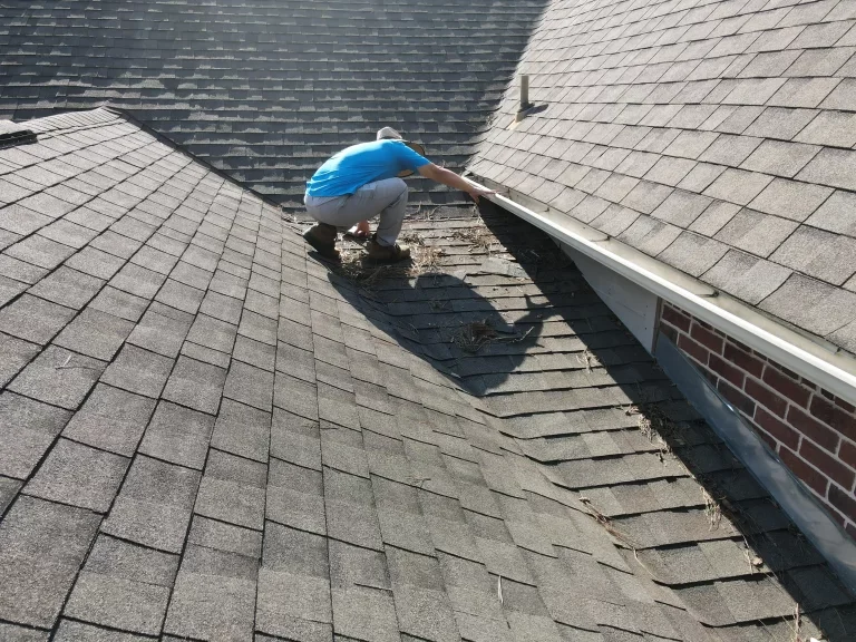 A person in a blue shirt and grey pants crouches on a shingled roof, cleaning debris from a gutter.