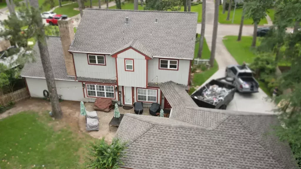 Aerial view of a house with gray siding, red trim, and gray roofs. A dumpster and truck are in the driveway.