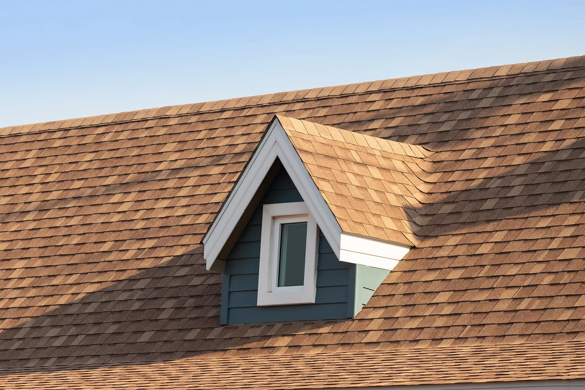 A dormer window with white trim, blue siding, and a gabled roof against a brown shingled roof and blue sky.