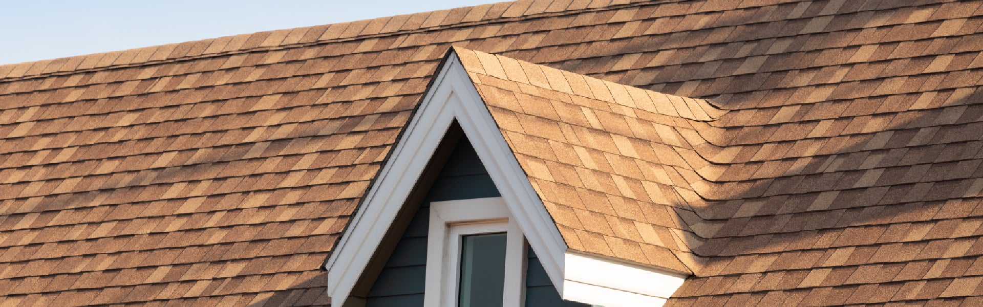 The roof of a house with weathered brown shingles, a dormer, and a window.