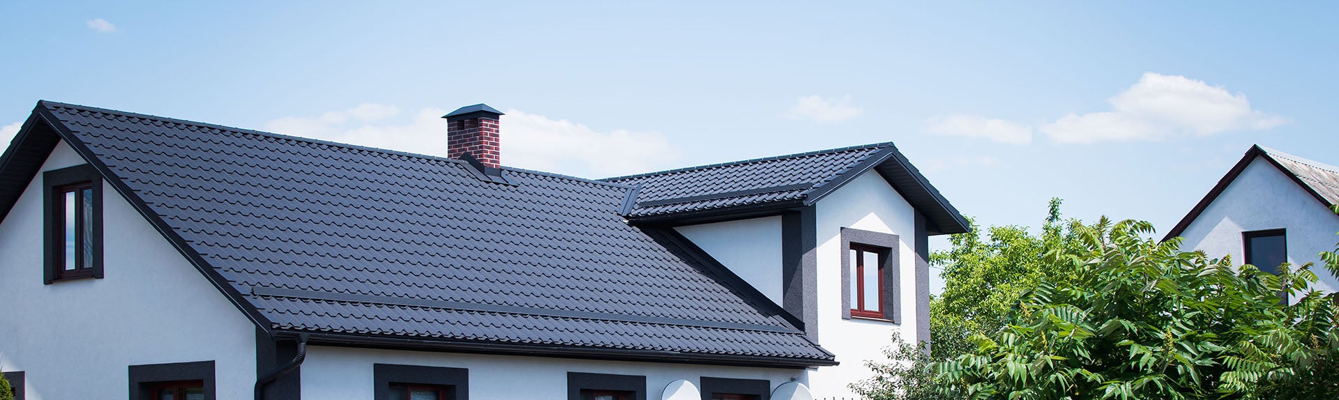 Modern white house with dark gray metal roof, brick chimney, dark trim, and green trees against a blue sky.