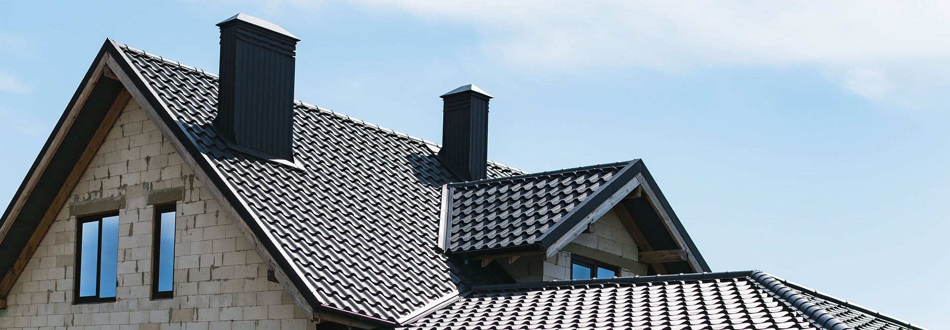 A textured, dark grey tiled roof with two chimneys and a light brick wall, against a clear sky.