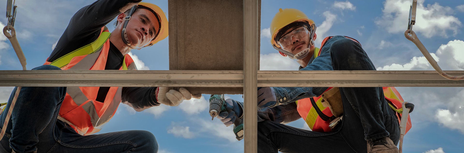 Two construction workers, viewed from below, install ceiling panels against a cloudy sky. Each wears a hard hat and safety vest.