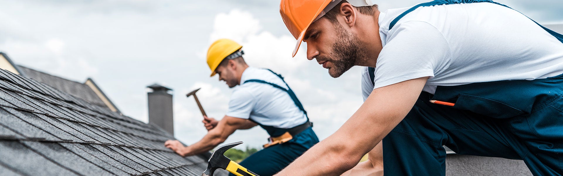Two roofers, wearing hard hats and overalls, repairing a shingled roof with hammers.