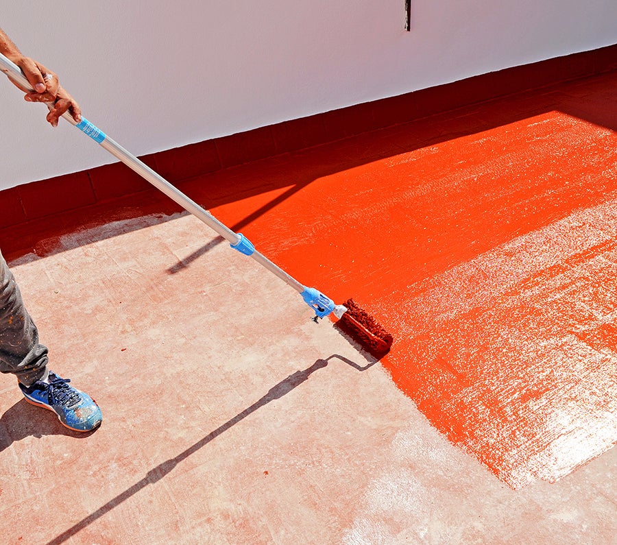 A person's hands and leg using a long-handled roller to apply bright orange paint to a concrete floor.