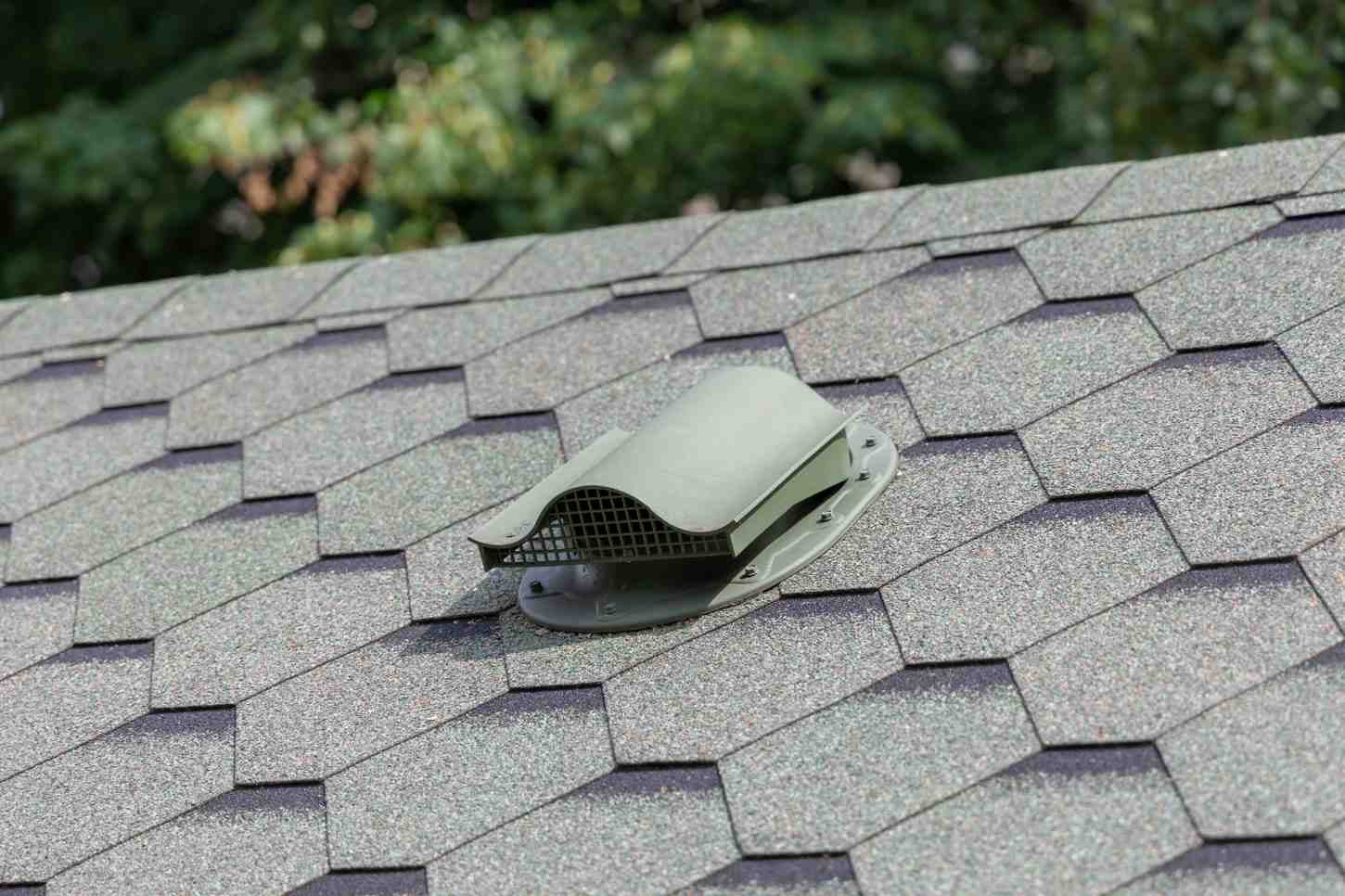 Close-up of a grey roof vent on a textured shingle roof, with blurred green trees in the background.