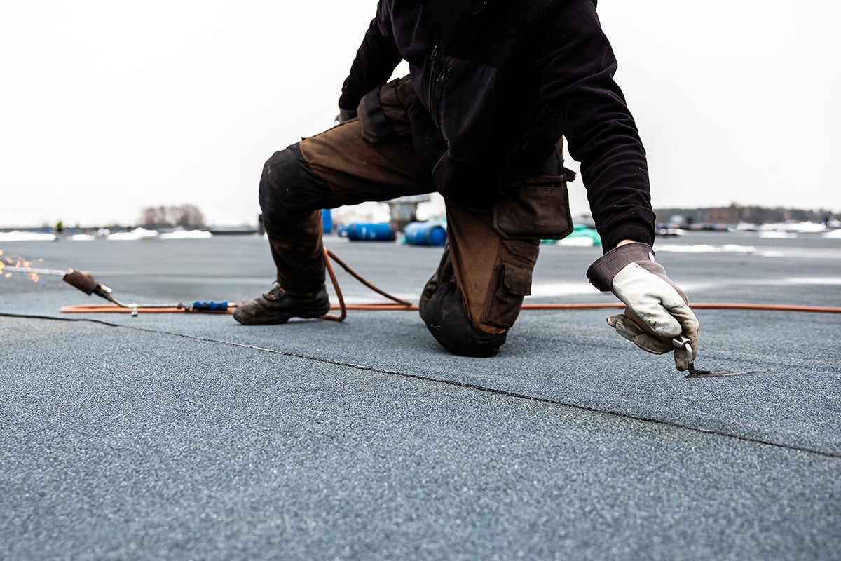 A worker in work pants and gloves kneels on a grey, textured roof surface, using a hook tool on a dark seam.