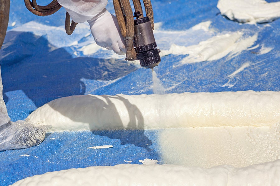 A person in protective gear sprays expanding white insulation foam onto a blue tarp, forming even layers.