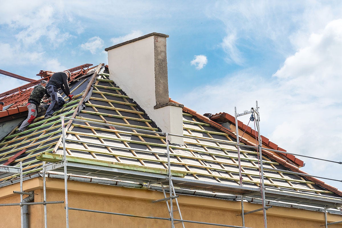 Two roofers work on a house roof under a blue sky, covered with black membrane, wooden battens, and some tiles.