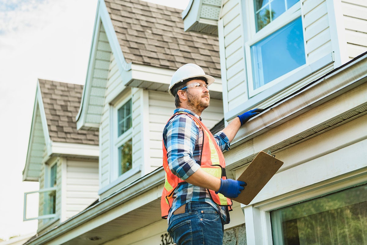 A home inspector in a hard hat and safety glasses examines the gutters of a house.