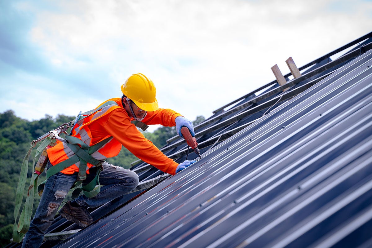A construction worker in a hard hat and safety harness installs solar panels on a slanted roof with a power drill.