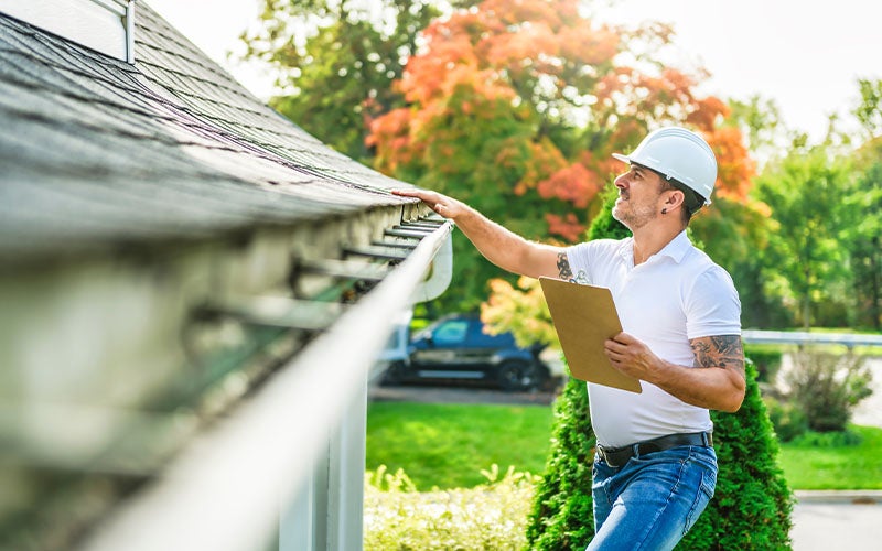 A man in a hard hat inspects a white gutter and gray roof shingles, holding a brown clipboard.